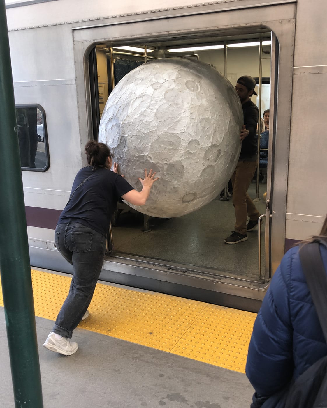 Two people struggle to push a huge handmade papier-mâché moon through the doorway of a commuter train.