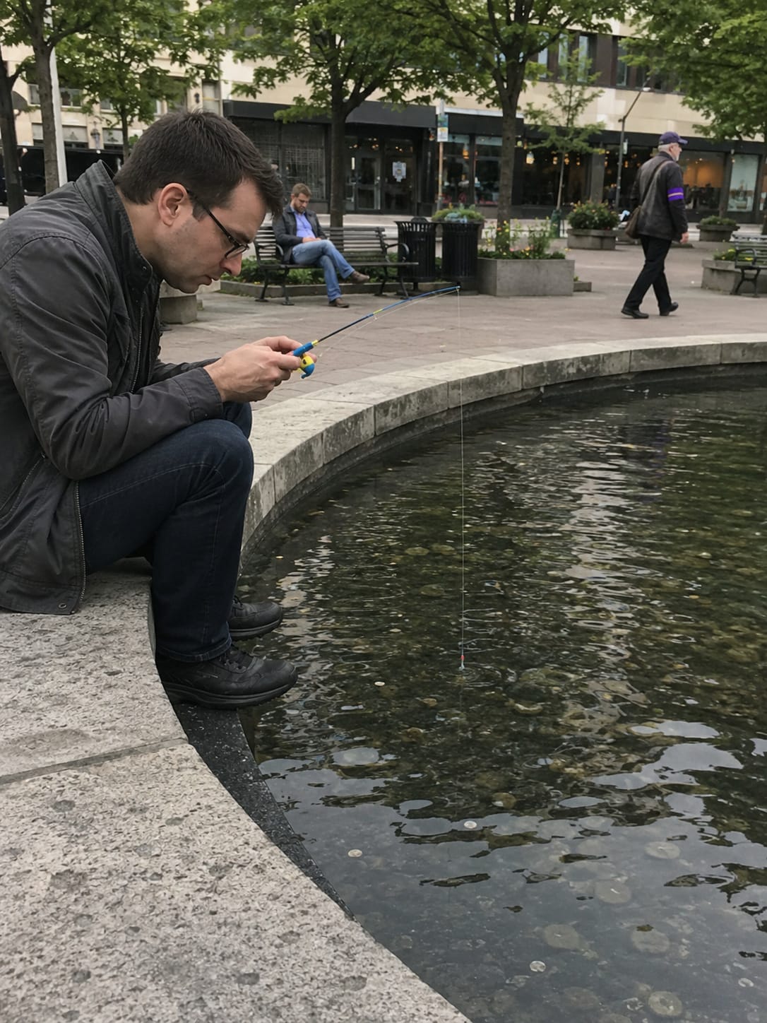 A serious man fishes with a tiny toy rod in a shallow public fountain filled with coins while pedestrians pass by.