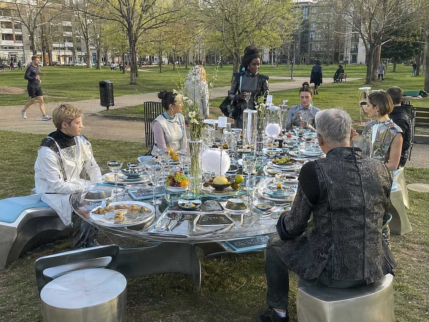 Futuristic diners in metallic clothing sit around a reflective high-tech banquet table in a modern park.
