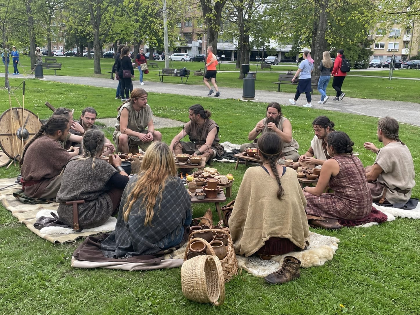 Iron Age diners in wool tunics share bread and bowls on blankets, with shields and spears beside them in a modern park.