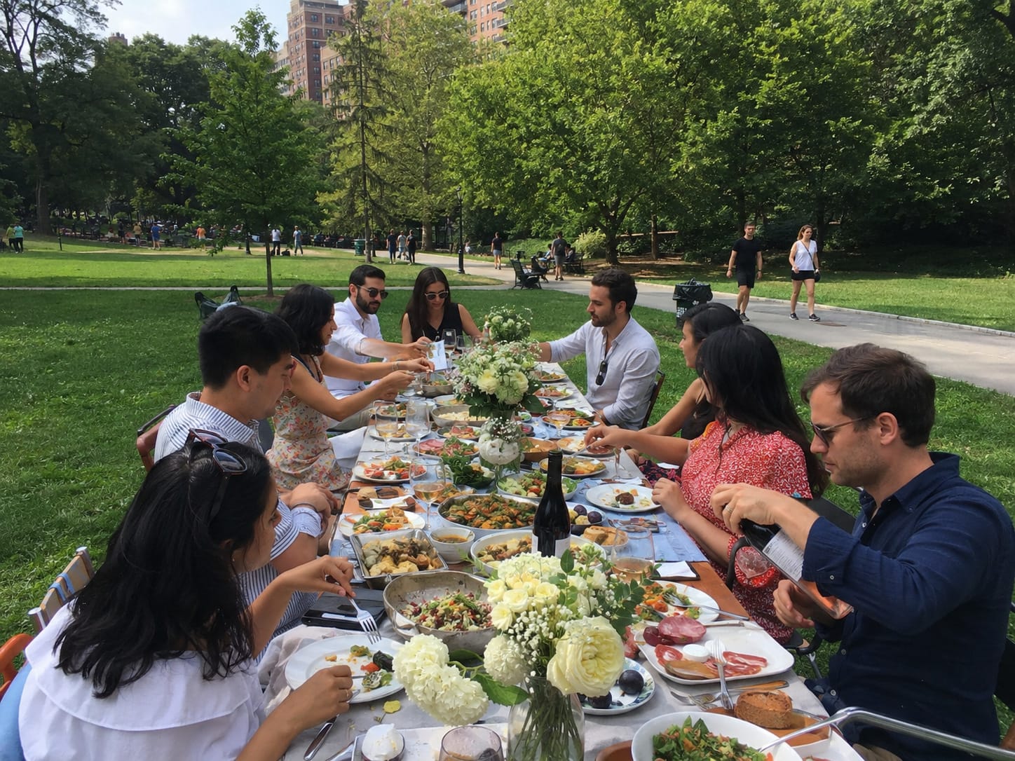 Contemporary guests share a lavish white-tablecloth picnic with flowers and wine in a sunny city park.