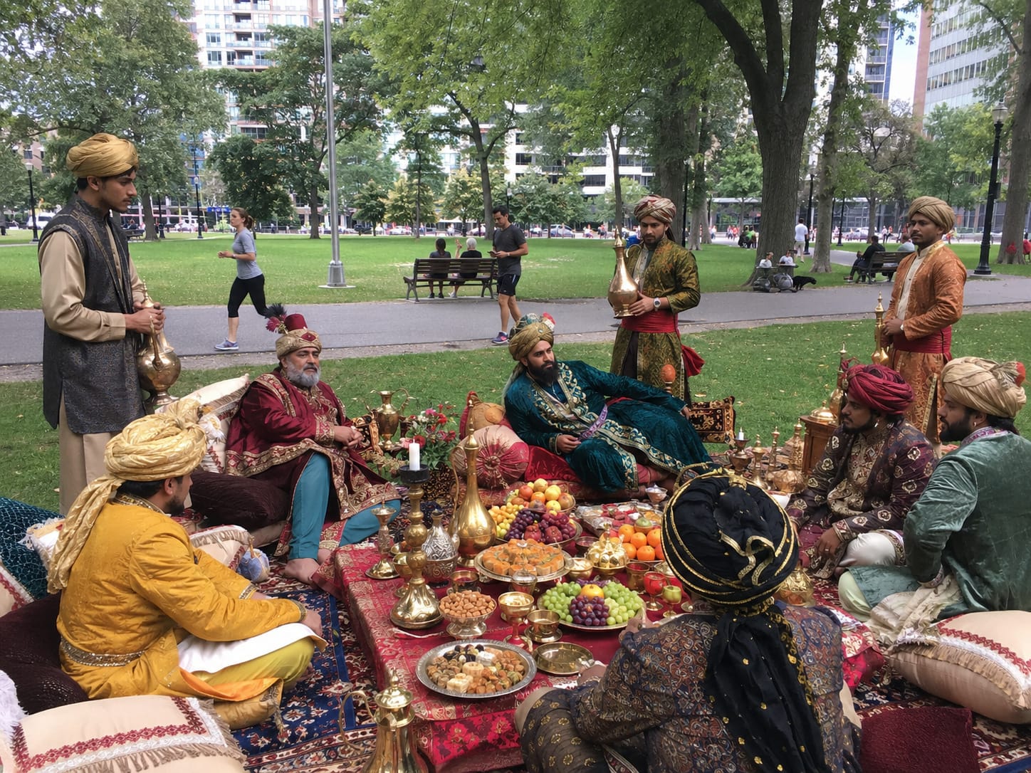 Mughal courtiers in jeweled robes and turbans dine on carpets amid brass vessels, fruit, and attendants in a modern park.