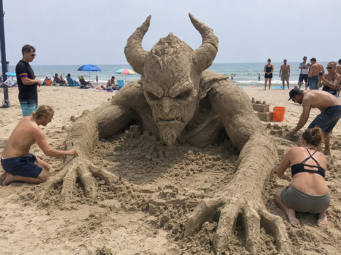 Giant demon sand sculpture appears to pull itself from the beach as visitors stand around it.