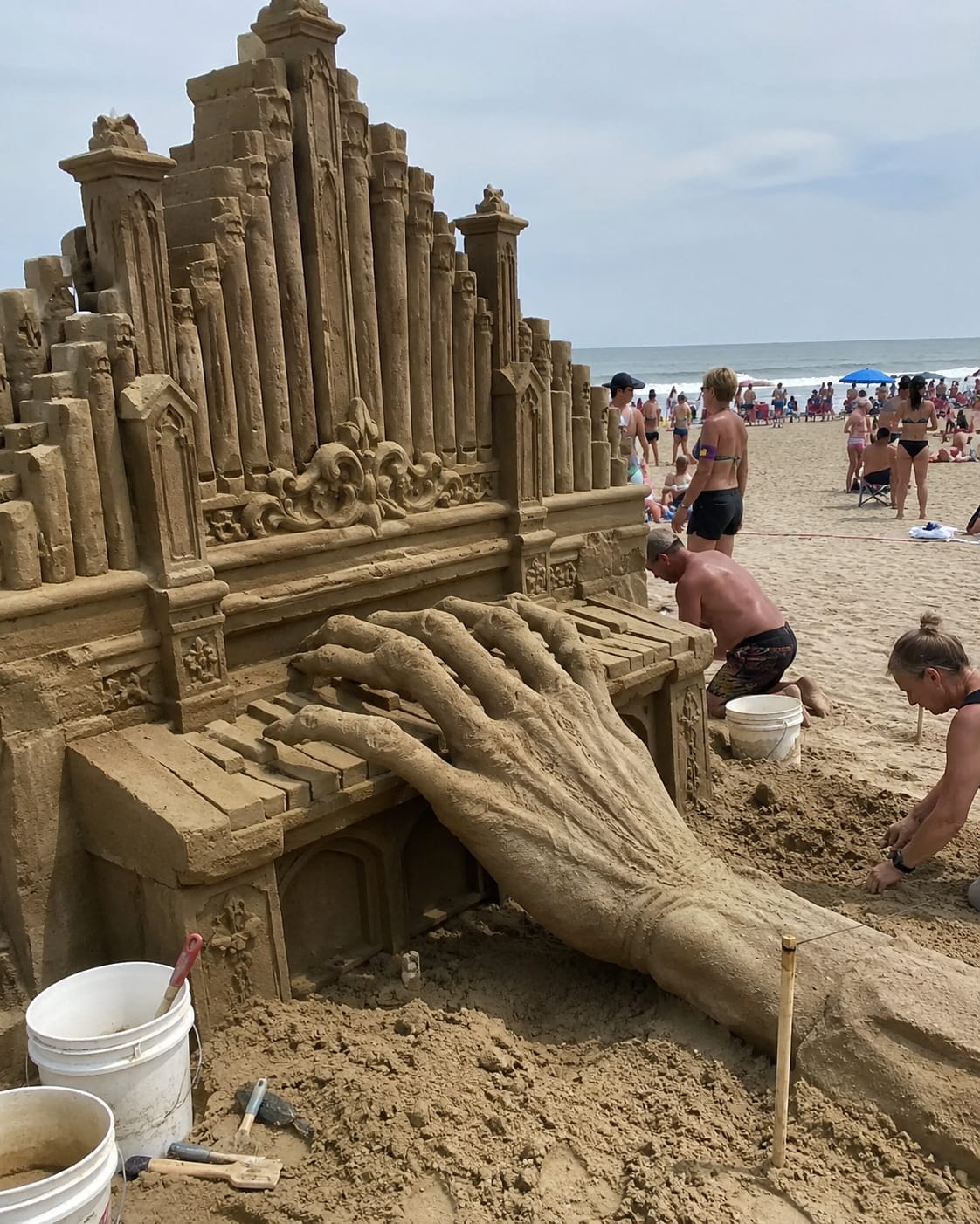 Massive sand pipe organ is played by an enormous many-fingered hand emerging from the beach.