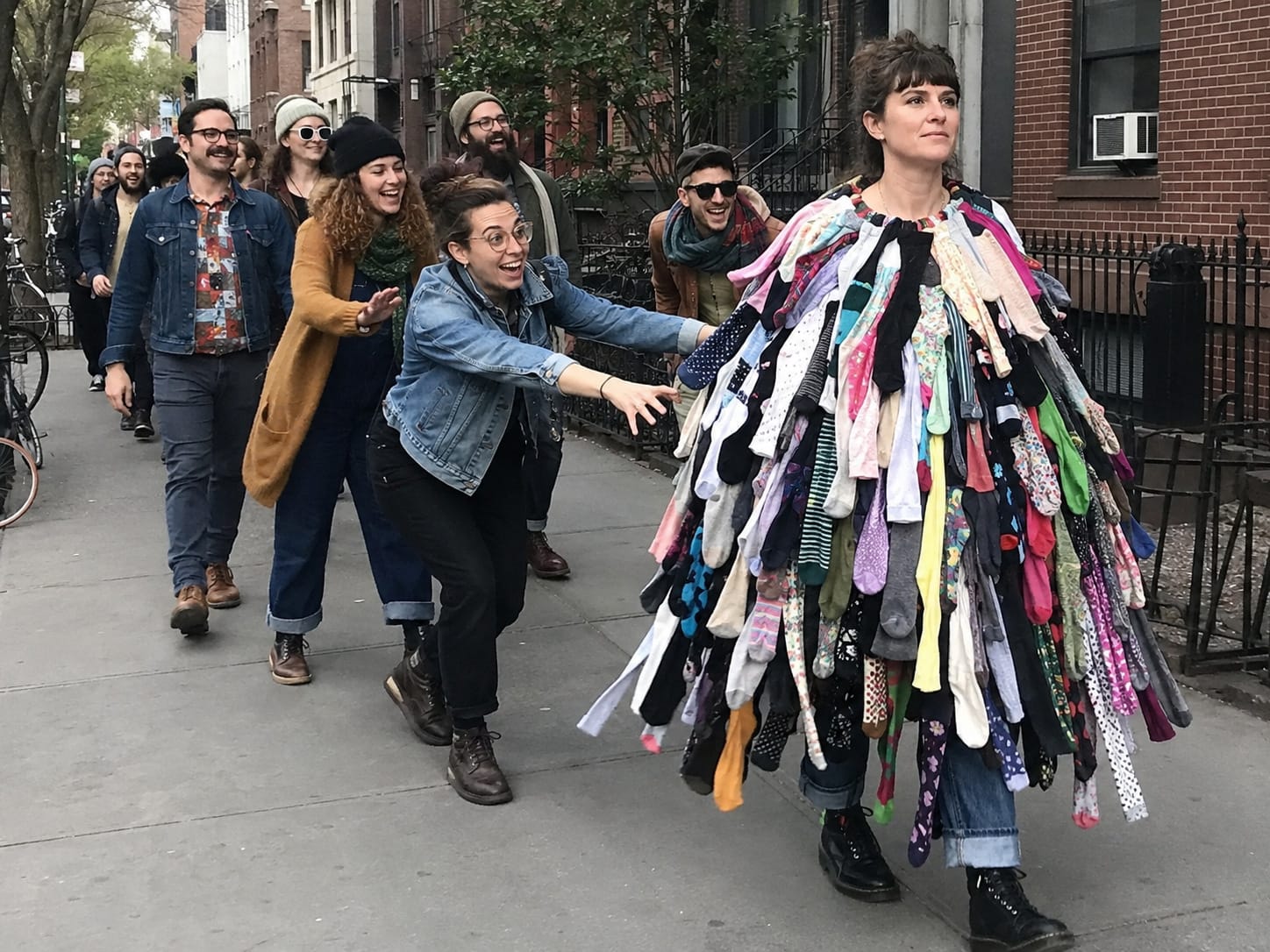 A woman in a cape of unmatched socks leads a laughing, dancing line of hipsters down a city sidewalk.