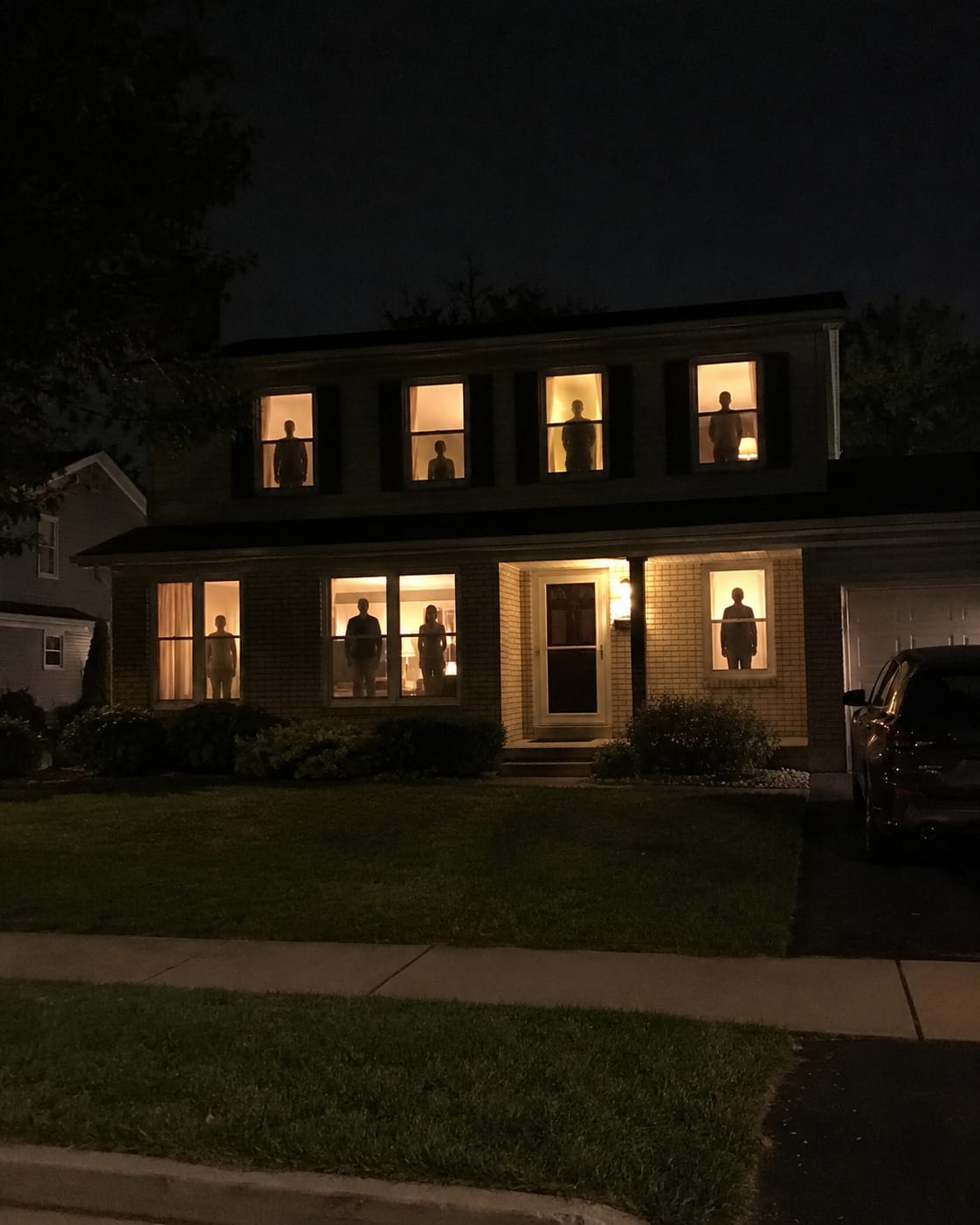 A suburban house at night, with a different human silhouette standing in every warmly lit window, watching the empty front yard.