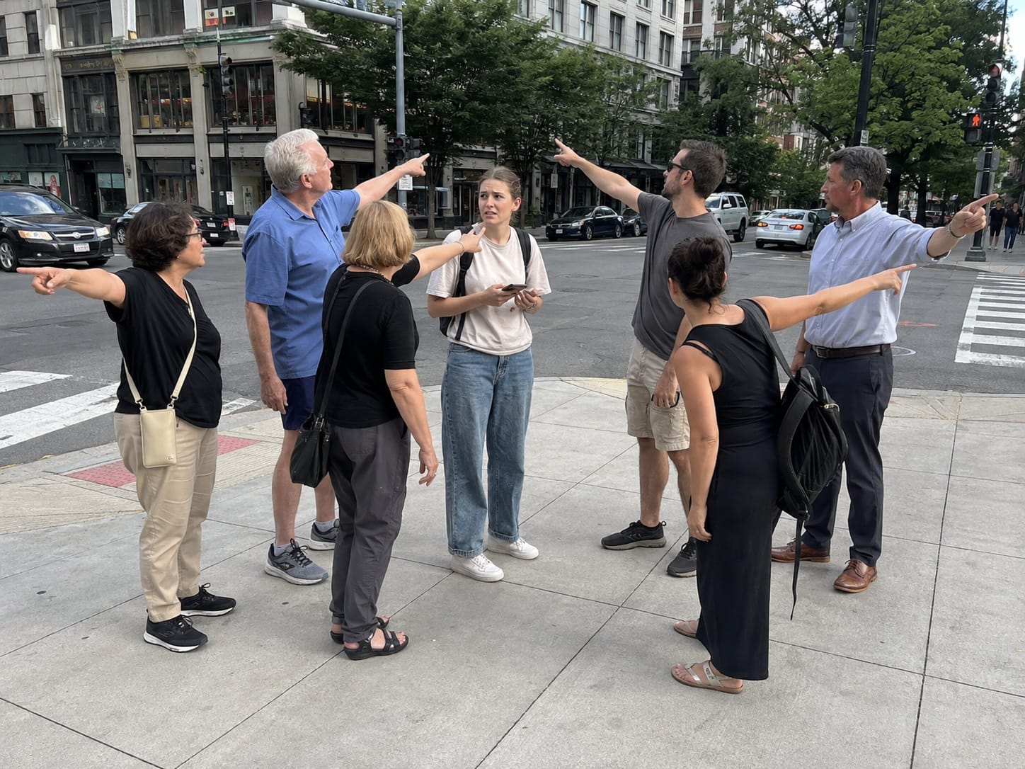 Confused tourist at a city intersection surrounded by six people simultaneously pointing in different directions.