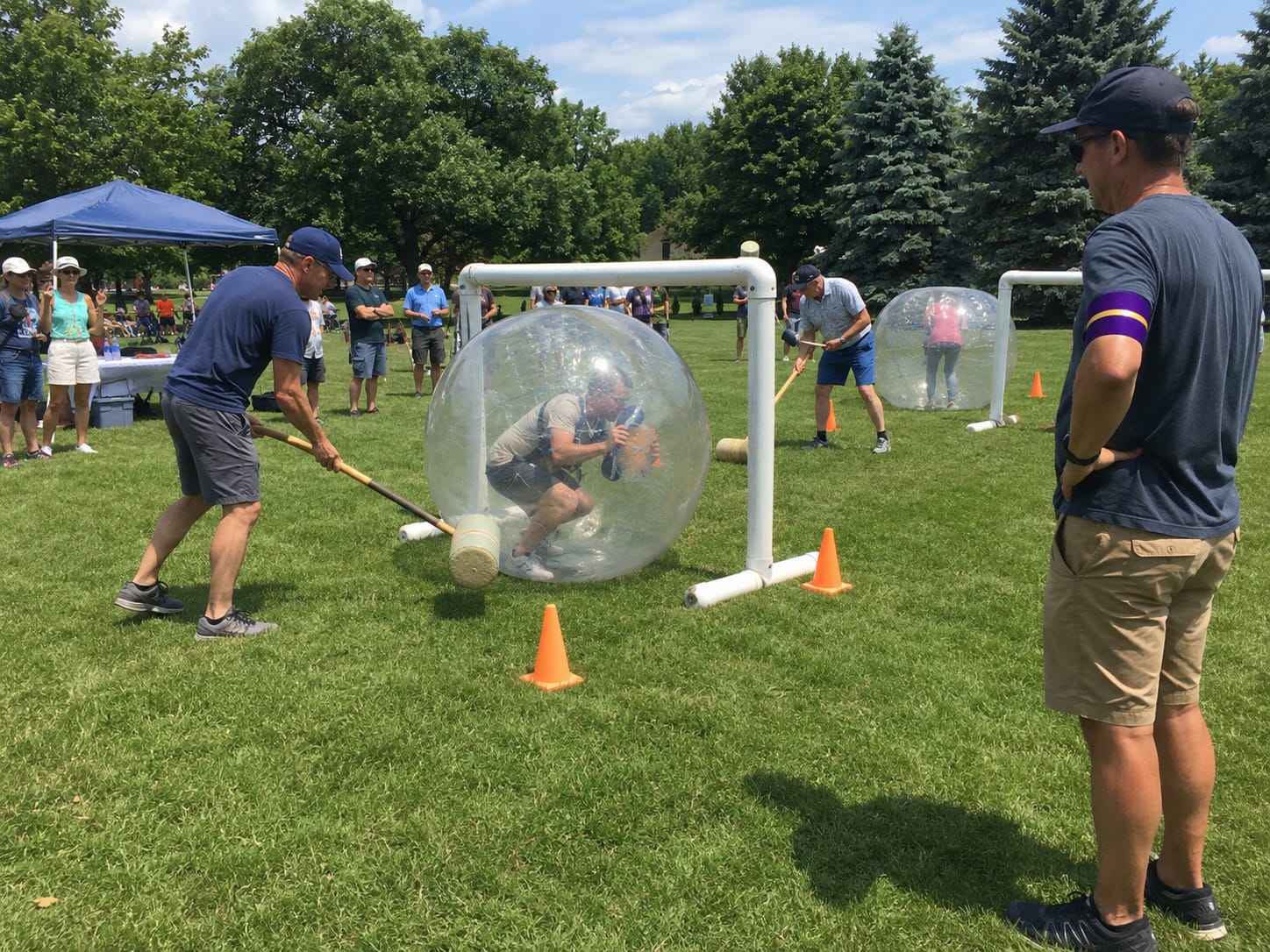 People inside giant clear inflatable balls are nudged through oversized croquet wickets on a grassy field.