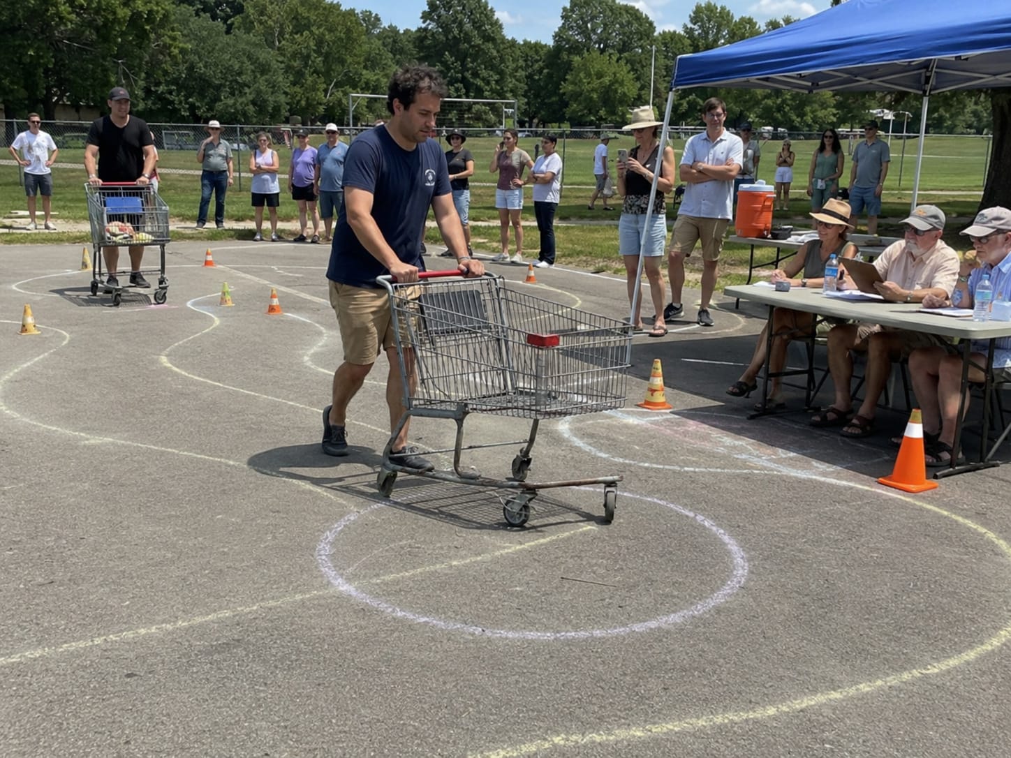 A competitor pushes a shopping cart through chalk curves and cones while judges watch from a folding table.