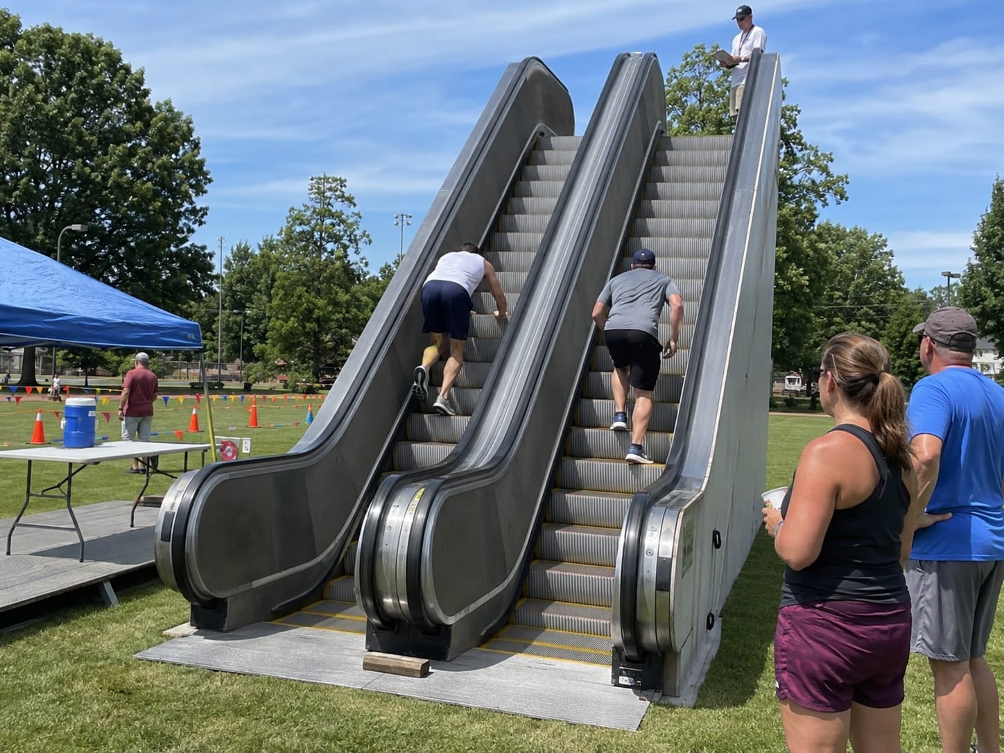 Two competitors climb side-by-side escalators set up in a park while spectators watch from the grass.