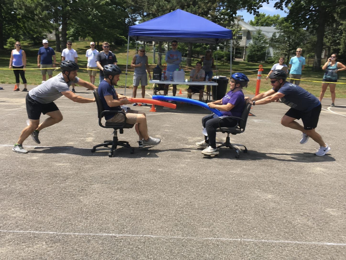 Two seated competitors roll toward each other in office chairs holding pool noodles while teammates push from behind.