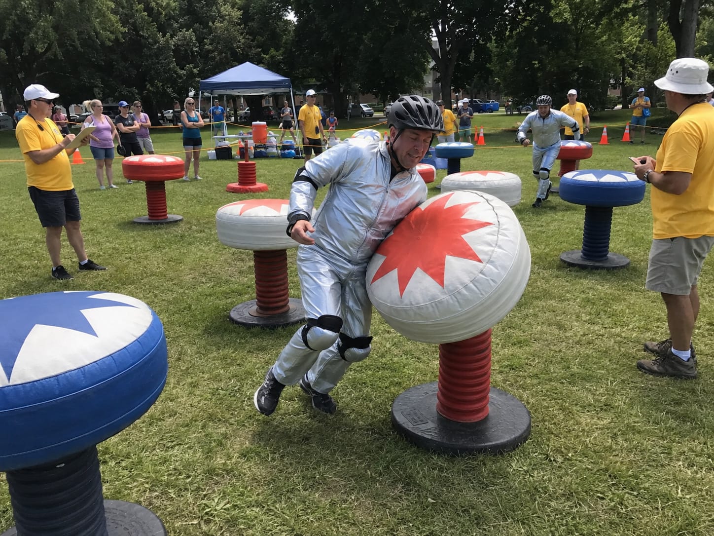 A competitor in a silver padded suit ricochets through a field of giant pinball bumpers as officials keep score.