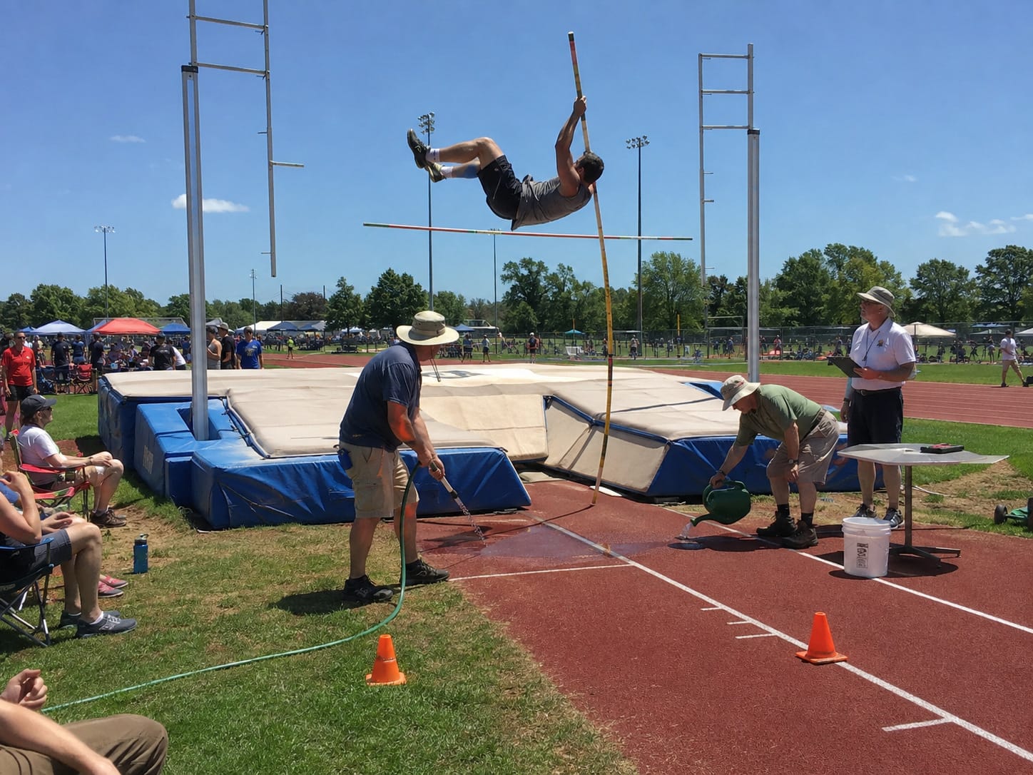 A pole vaulter sails over a tiny puddle while two officials on the ground keep refilling it with hose and watering can.