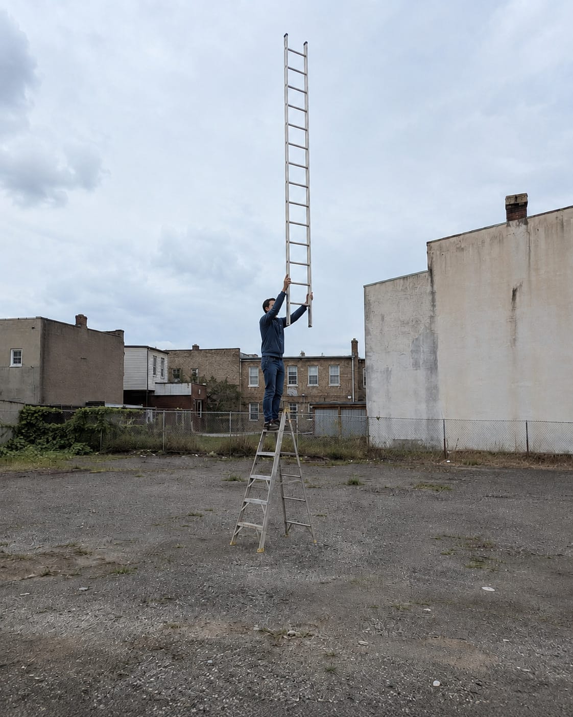 Man stands on a stepladder in an empty lot, holding a second full-size ladder vertically above his head.