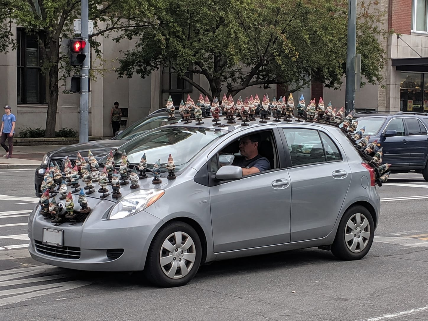 Compact hatchback stopped at a red light, covered with dozens of forward-facing garden gnomes across the hood, roof, and rear.
