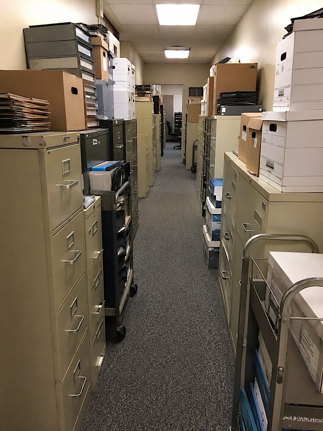 A fluorescent-lit office hallway crowded on both sides with filing cabinets, banker boxes, carts, binders, and old paperwork, leaving only a narrow path down the middle.