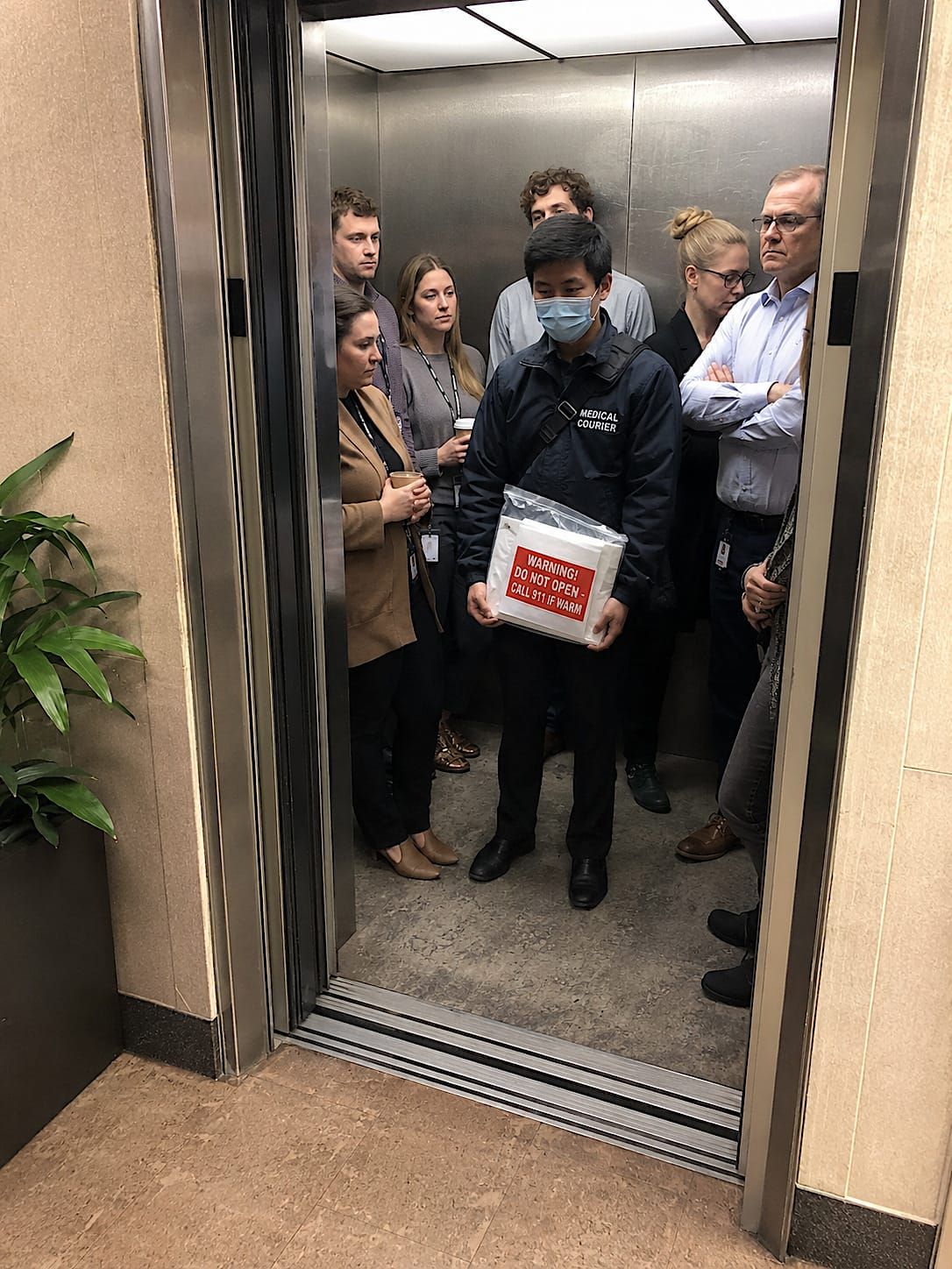 A crowded office elevator seen from the lobby, with tense office workers surrounding a masked medical courier holding a warning-labeled transport package.