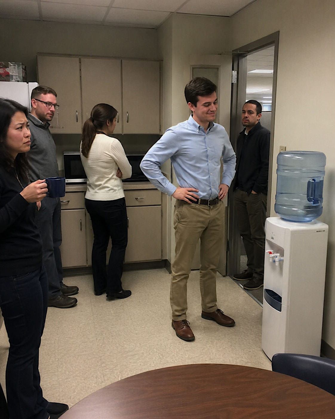 Young intern standing proudly beside an office water cooler while coworkers react with concern.