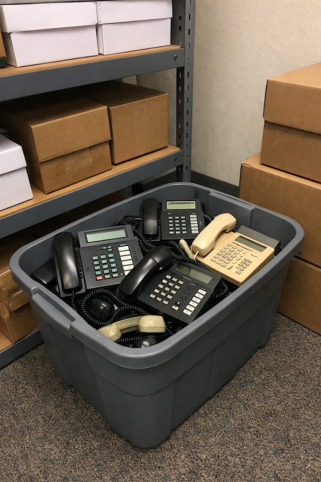 Plastic storage bin filled with old office telephones, tangled cords, and mismatched handsets.