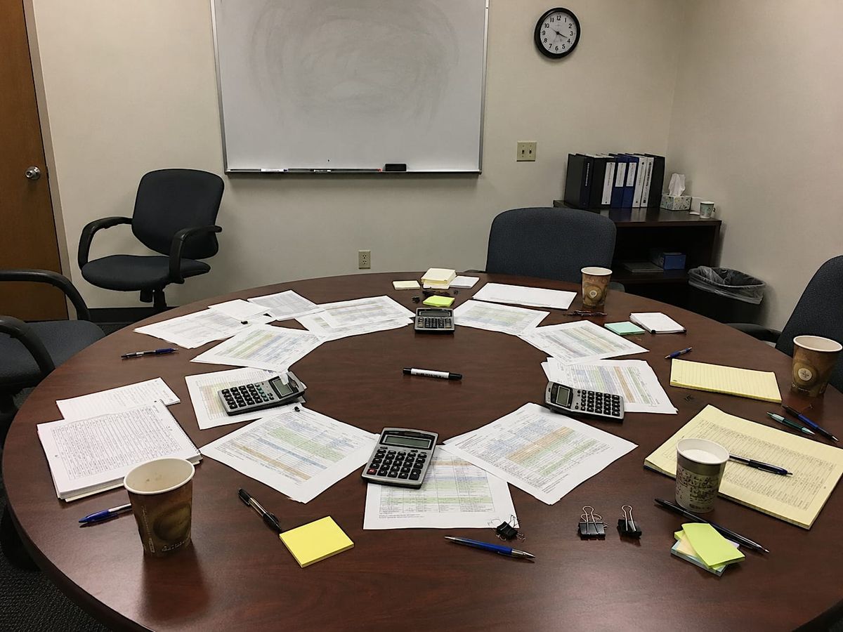 Spreadsheet pages, calculators, coffee cups, and a dead marker form a ritual-like circle on an office conference table.