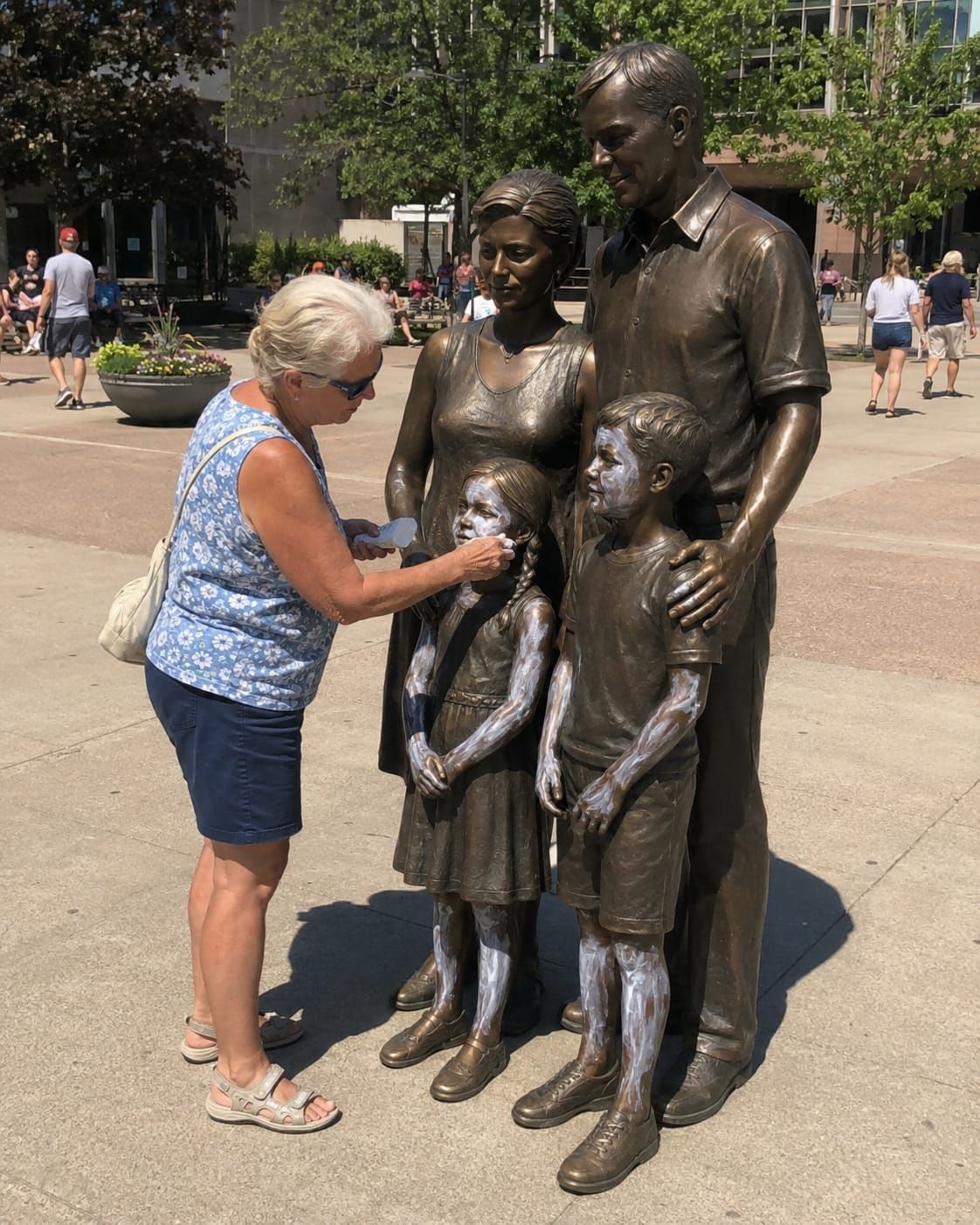 Older woman applies sunscreen to the child figures of a bronze family sculpture in a sunny public square.