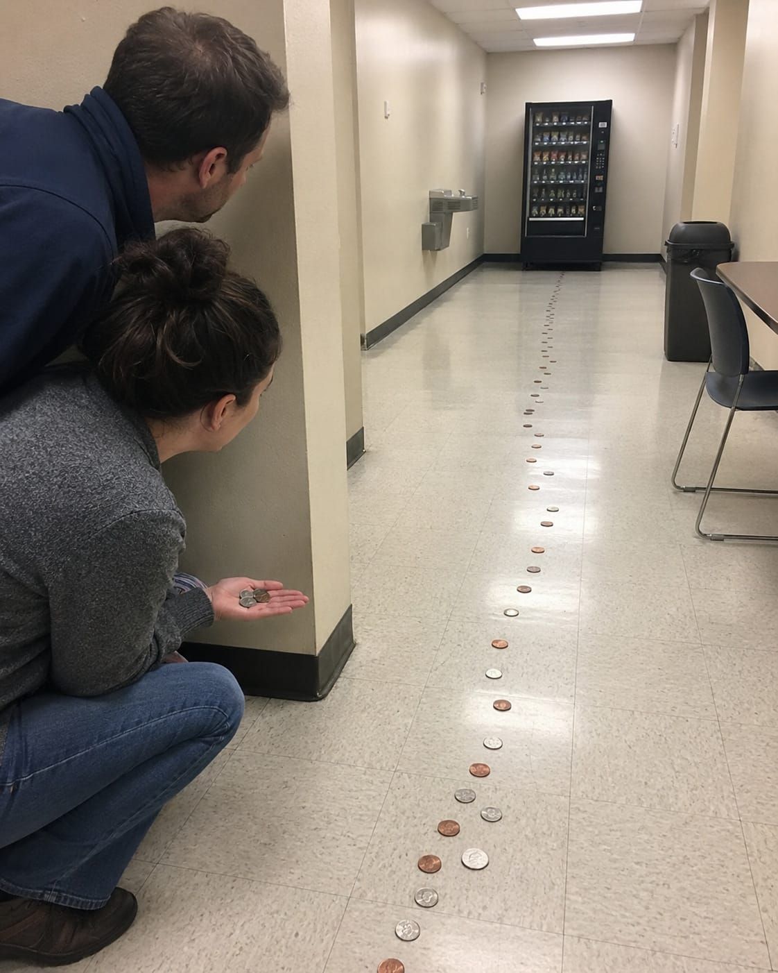 Two adults hide around a corner while a coin trail leads to a vending machine that refuses to move.