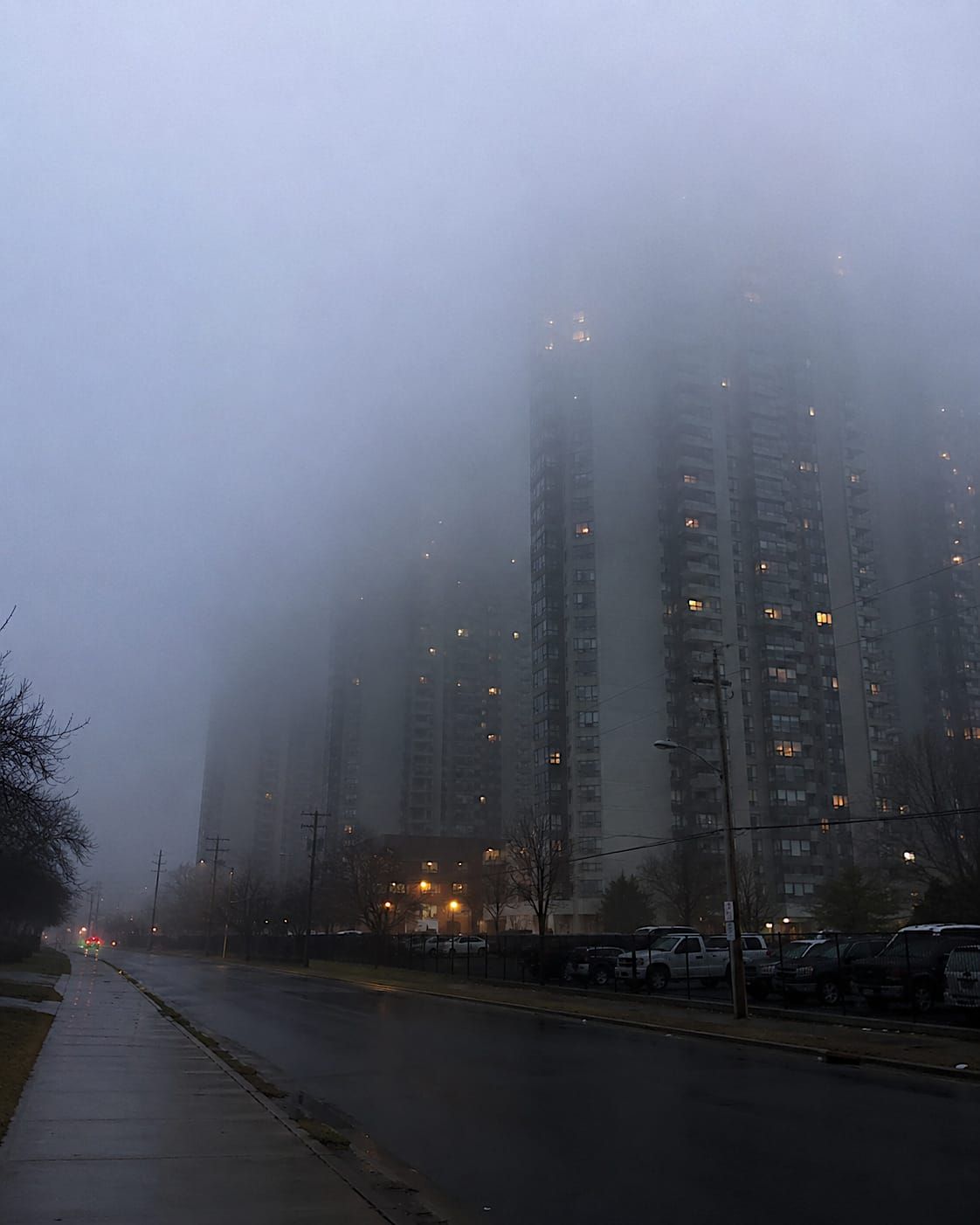Apartment towers rise through early morning fog above a wet residential street, their upper floors fading into low cloud while a few warm windows glow in the gray dawn.