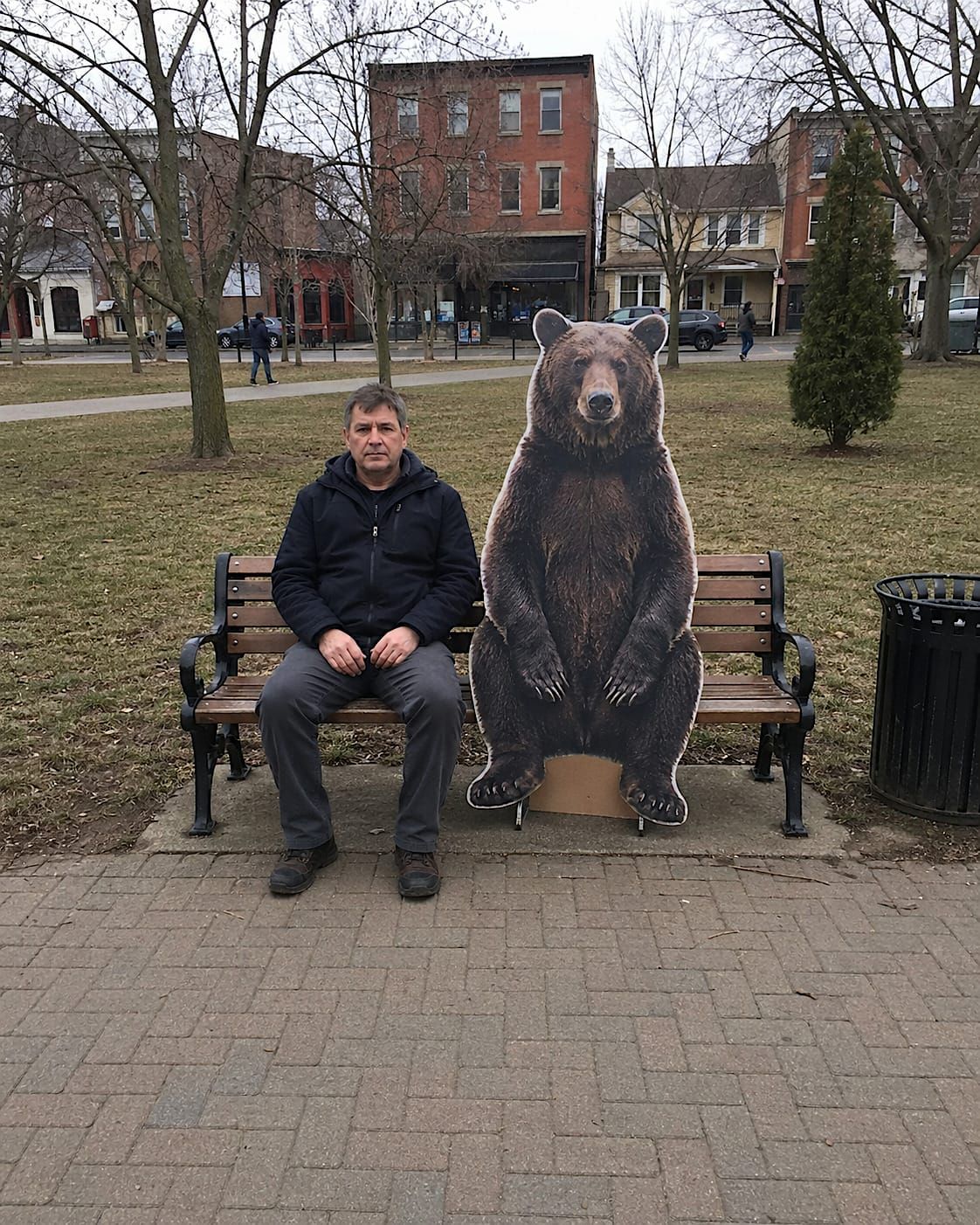 Ordinary man sitting on a park bench beside a life-size cardboard cutout of a bear.