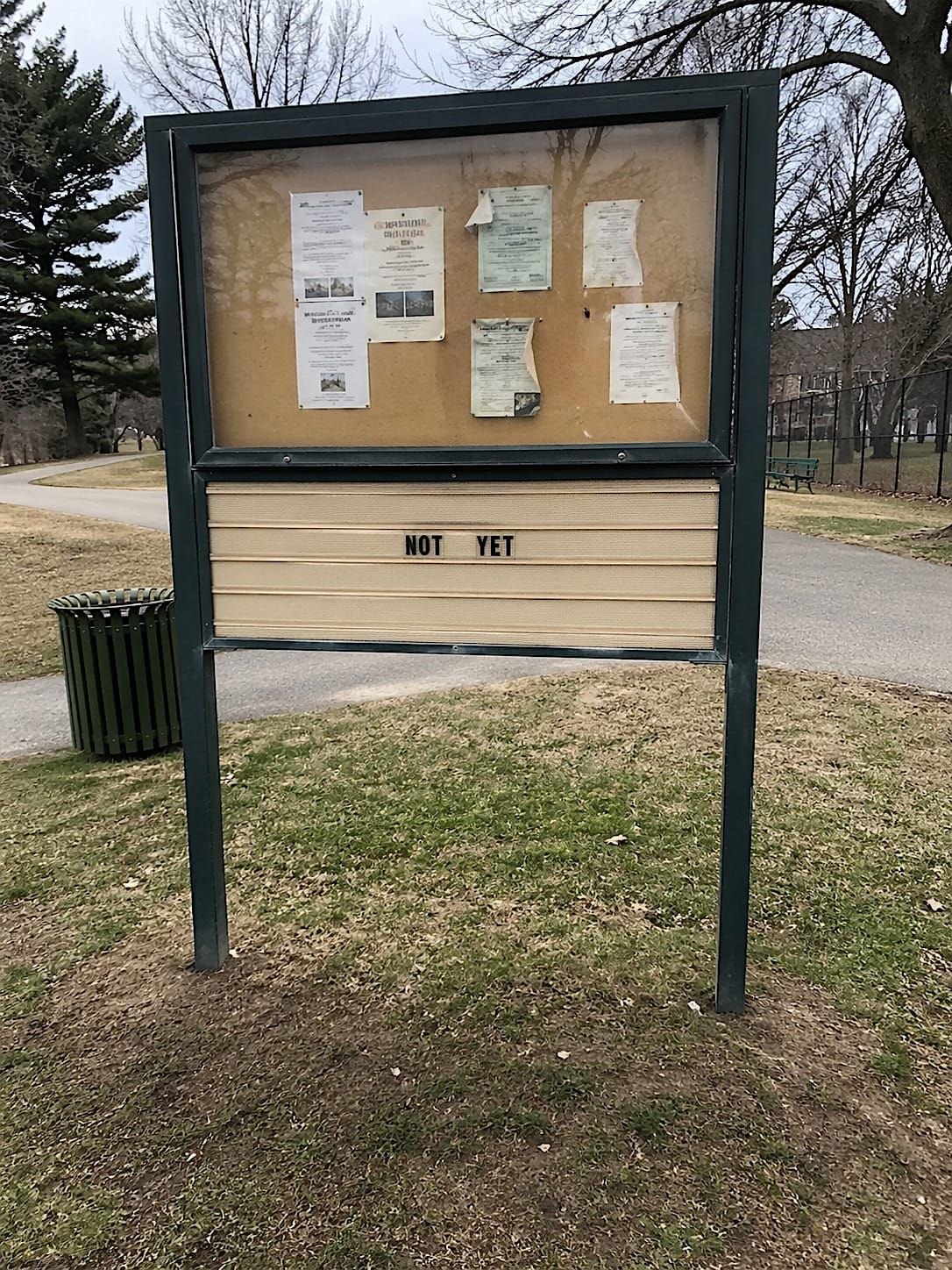 Community notice board in a neighborhood park with faded flyers and a letter panel reading “NOT YET.”