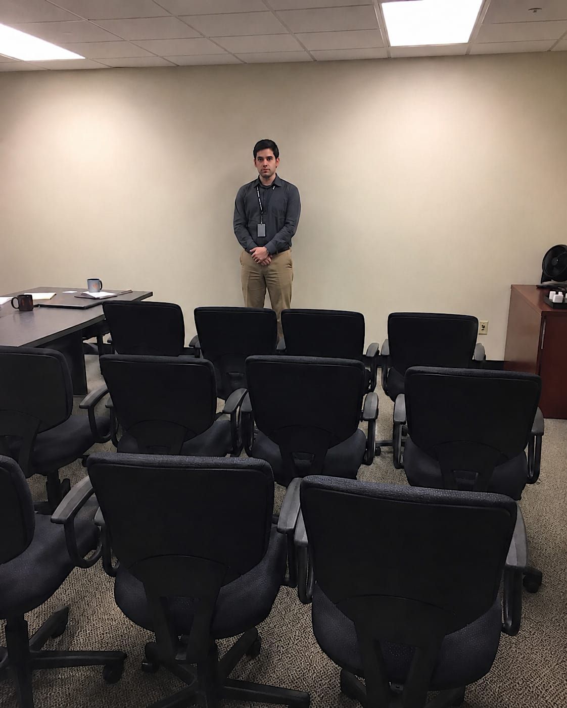 Rows of rolling office chairs facing one uncomfortable employee in a plain conference room.