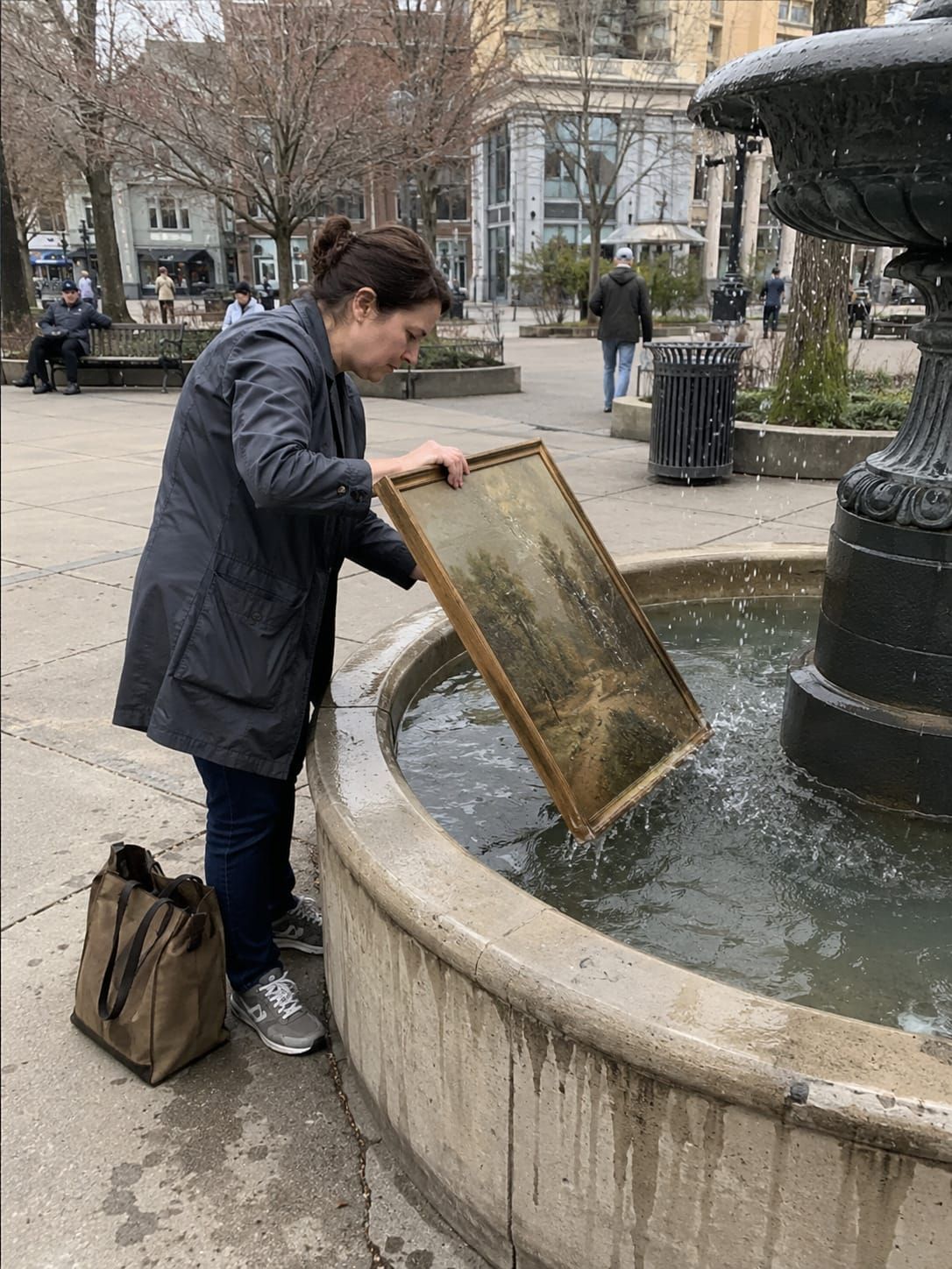 A woman carefully rinses a large framed painting in a public fountain while passersby mostly ignore her.