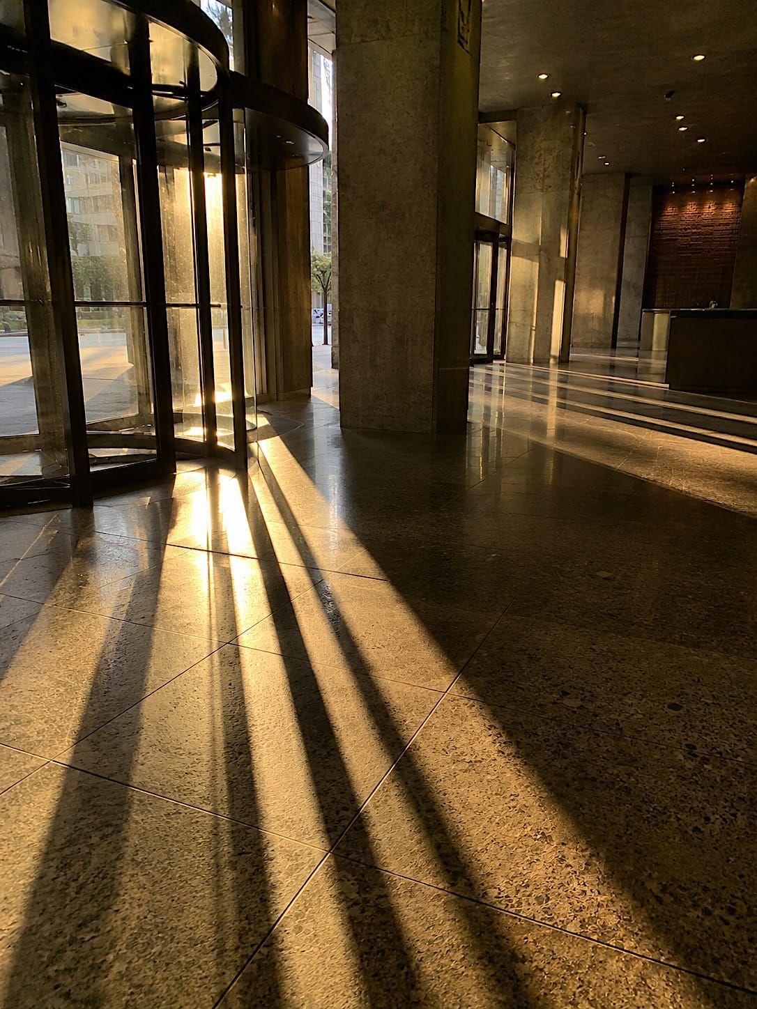 Golden sunlight casts long revolving-door shadows across a polished brutalist office lobby floor.