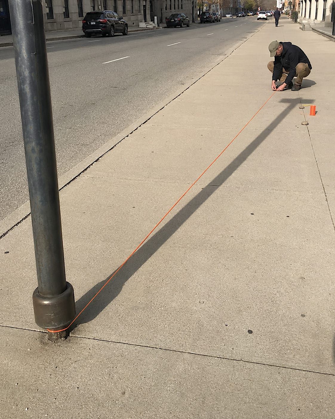 An ordinary person crouches on a city sidewalk, carefully measuring a lamppost’s long shadow with bright orange surveyor’s string.
