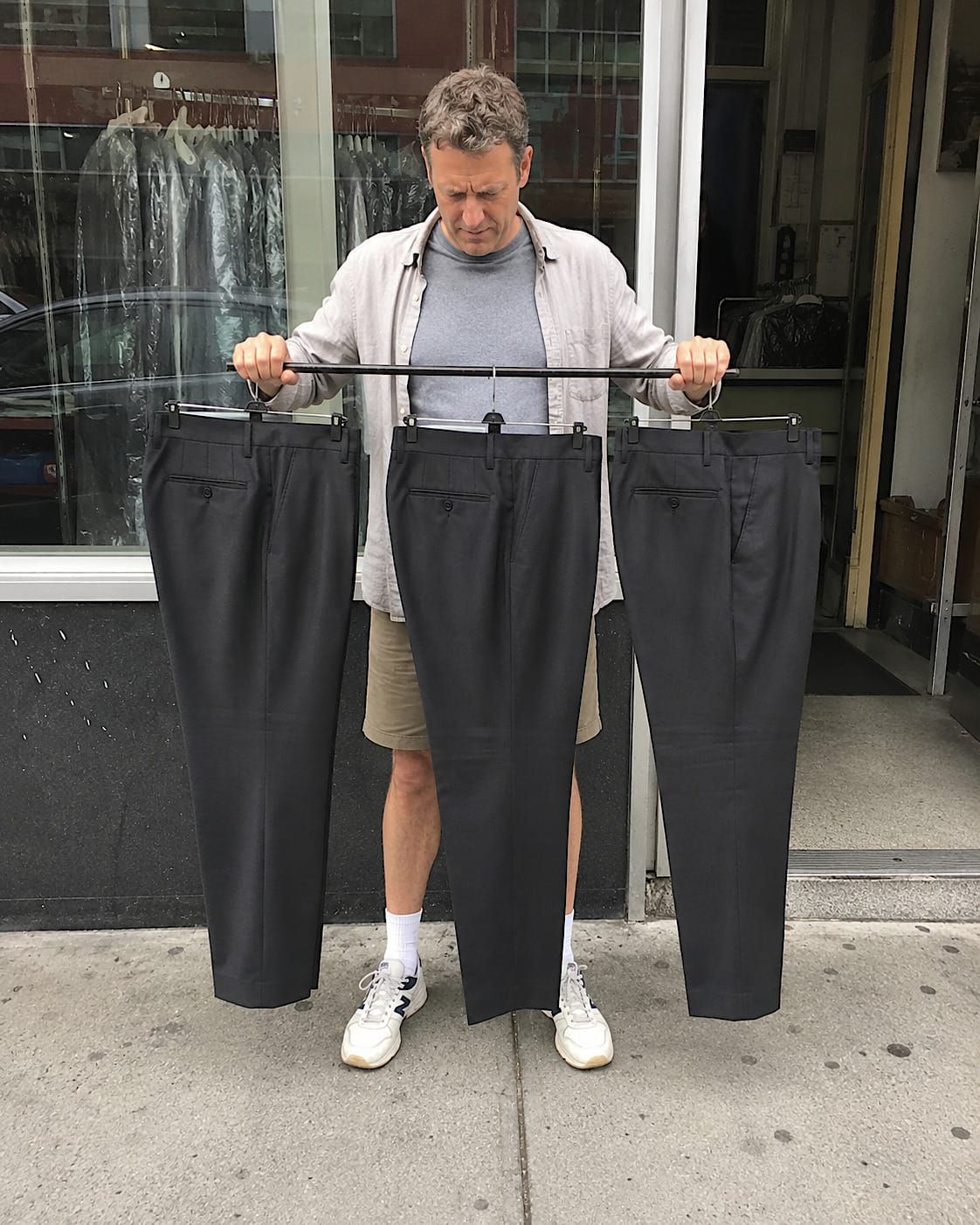 Man in shorts studying three pairs of dark dress pants on hangers outside a dry cleaner.