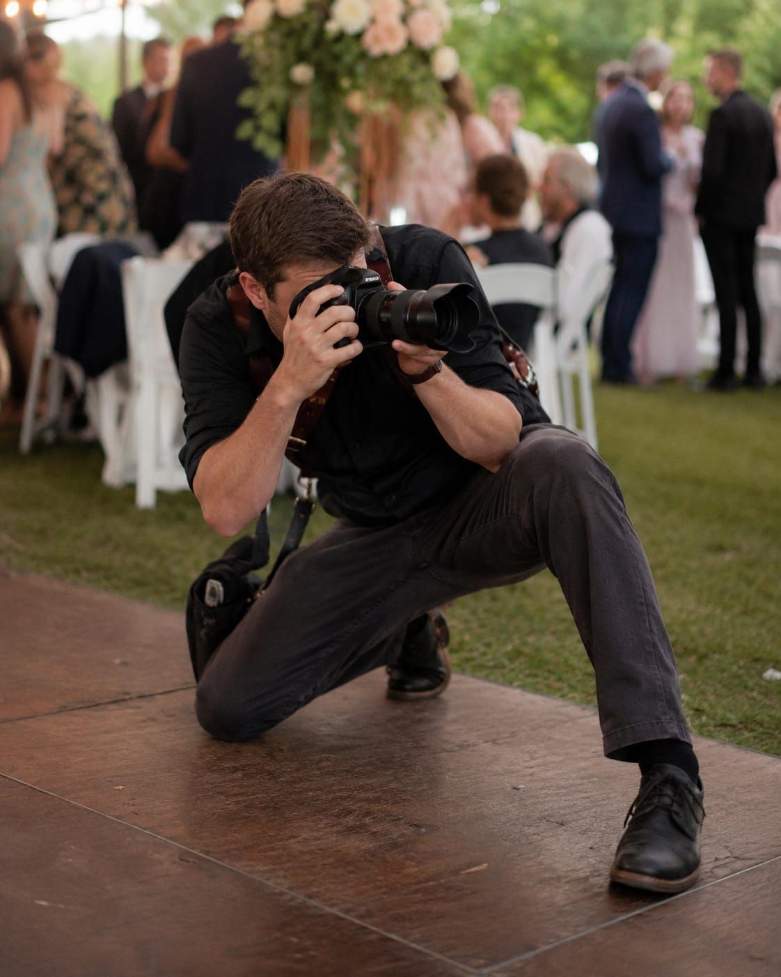 Wedding photographer crouches awkwardly for a shot while dressed-up guests blur behind him at a modest outdoor reception.