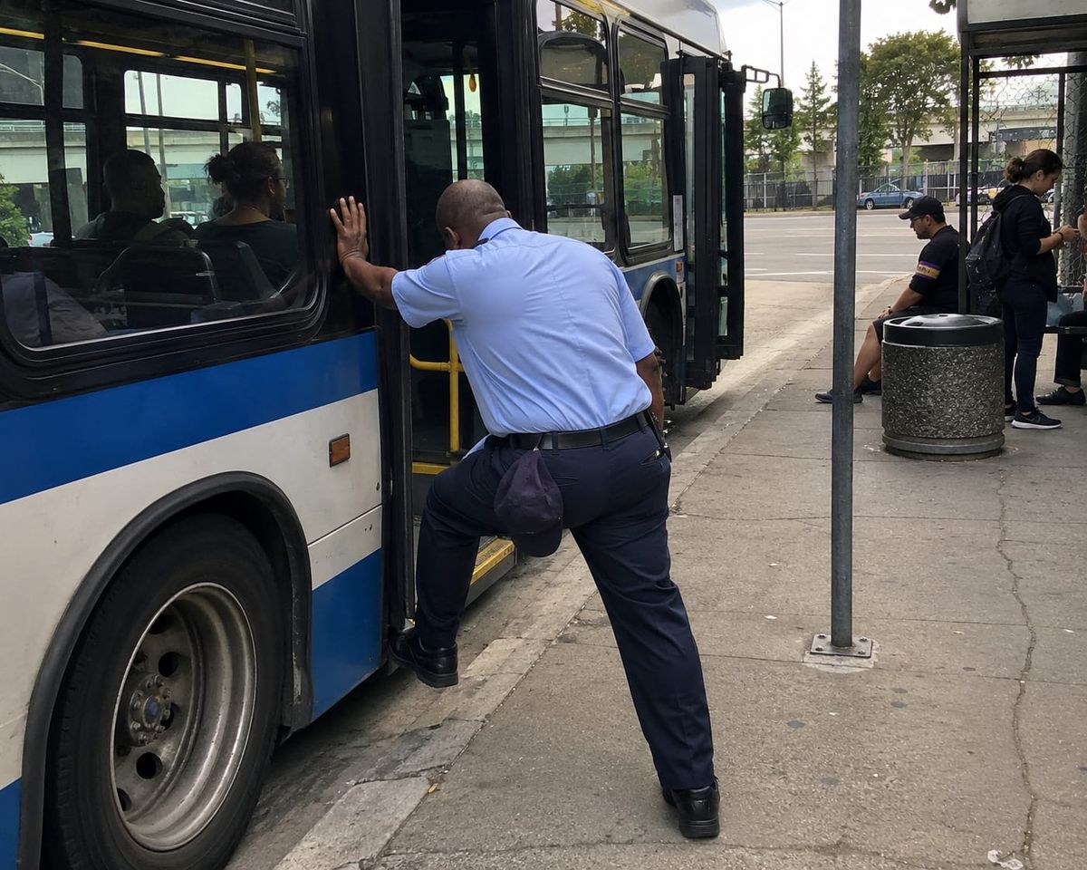Uniformed bus driver stretches beside a stopped city bus during a layover while passengers wait nearby at the curb.