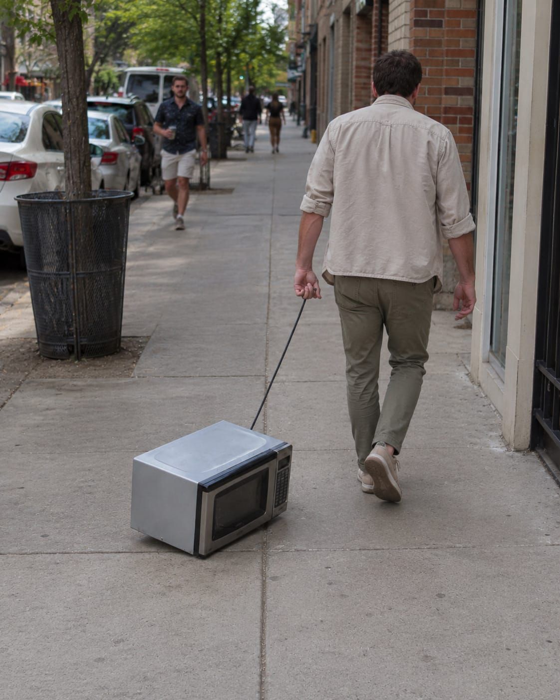 A man drags a microwave down the sidewalk by its cord like a stubborn pet refusing to keep walking.