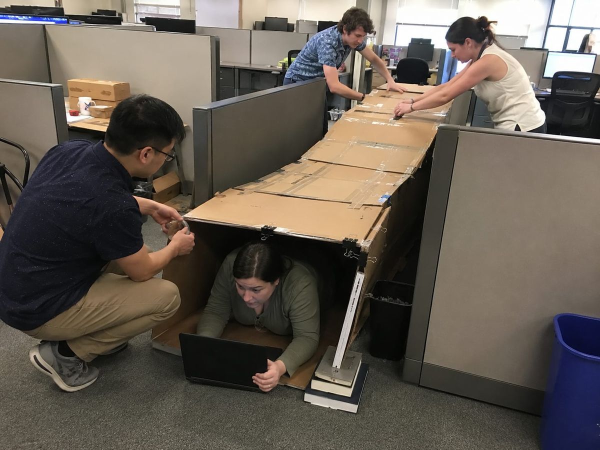 Coworkers build a cardboard tunnel between cubicles while a woman crawls through it holding a laptop.