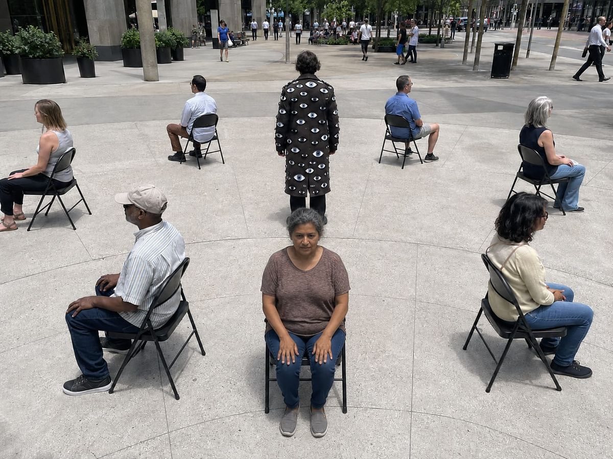 Public performers sit facing away from a central figure wearing a dark coat covered in sewn-on fabric eyes.