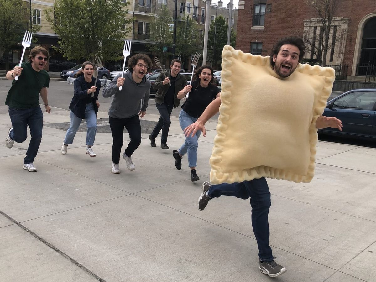 A man in a giant ravioli costume runs down a city sidewalk as a laughing crowd chases him with oversized forks.