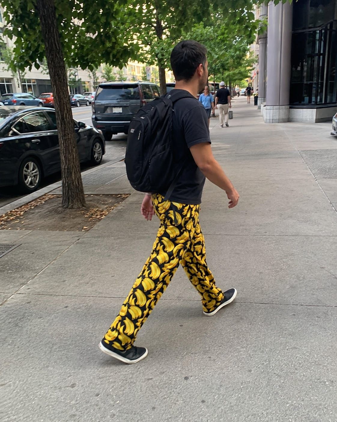 Man walking on a city sidewalk in black pants densely patterned with bright yellow bananas.