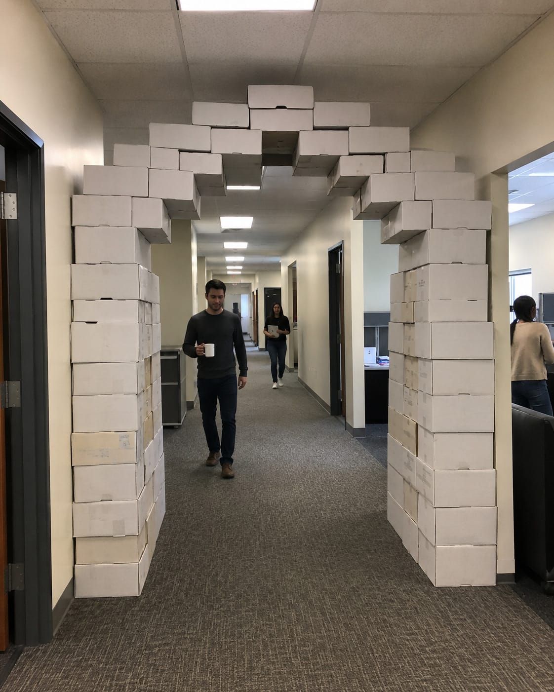 Printer-paper boxes form a full office archway as employees casually walk beneath it in a bland fluorescent corridor.