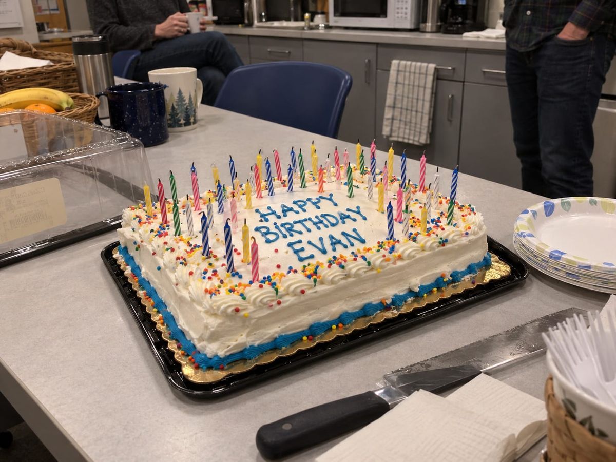 Office birthday cake reading “Happy Birthday Evan,” covered in candles, with coworkers gathered around a break-room table.