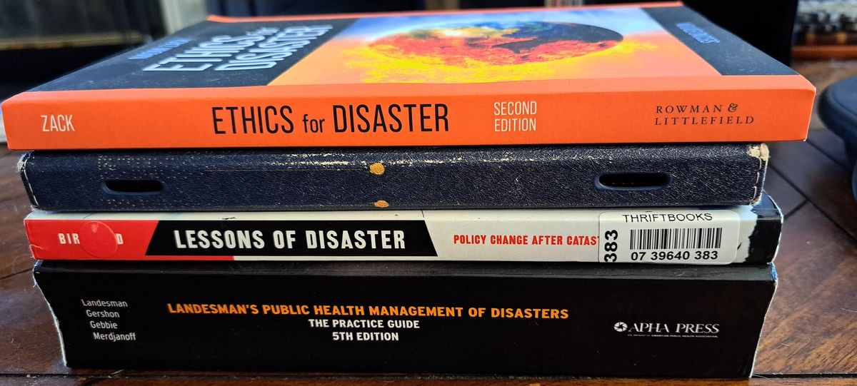A stack of books on a wooden coffee table: Landesman's Public Health Management of Disasters, Birkland's Lessons of Disaster, a Kindle Fire, and Zack's Ethics for Disaster