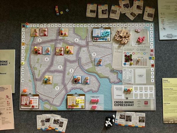 View from above at the board game Cross Bronx Expressway. Wooden pieces in multiple colors, cards detailing historical events, and tokens to track board state. 