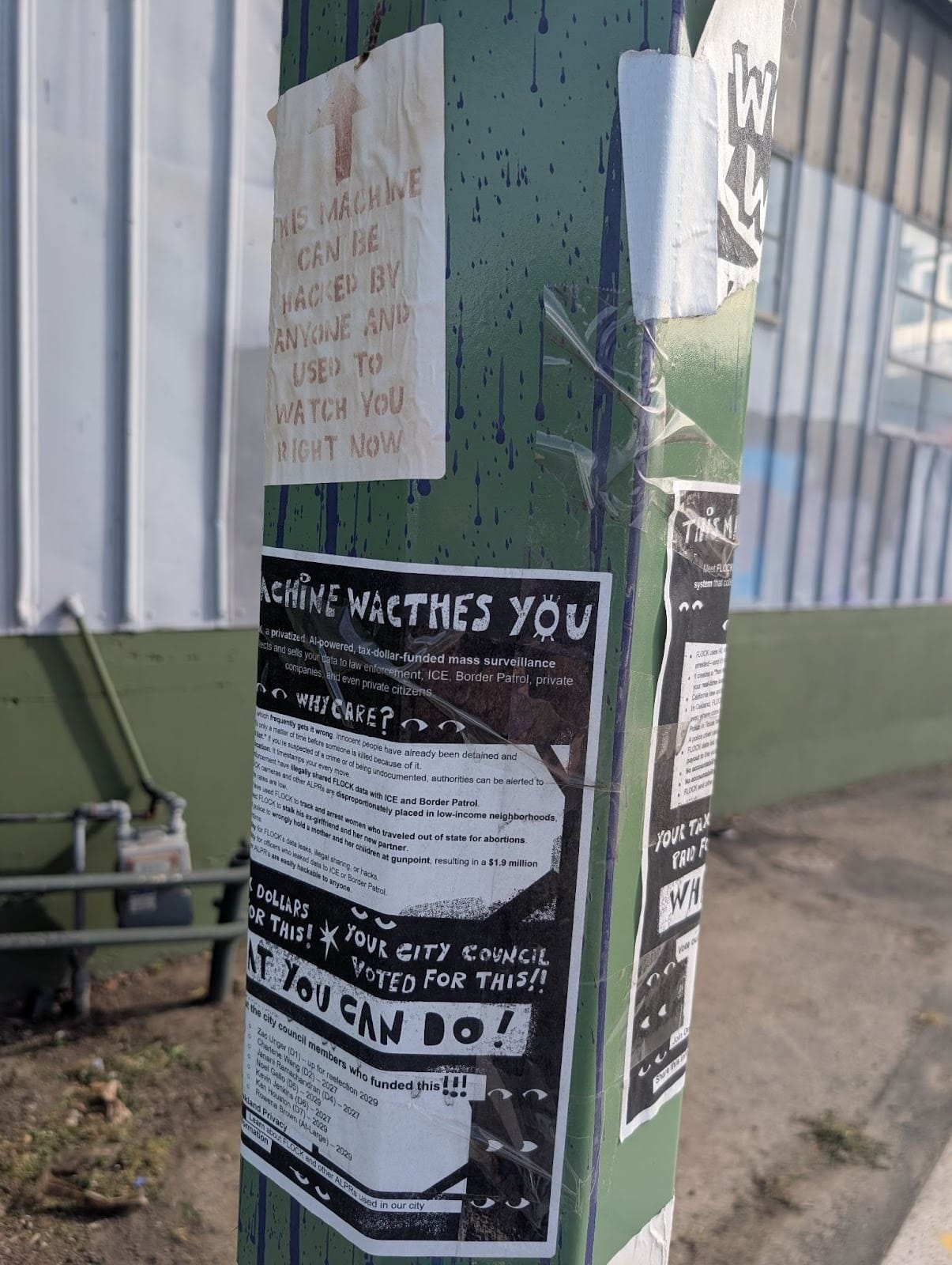 Two signs on a post. One reads, "THIS MACHINE CAN BE HACKED BY ANYONE AND USED TO WATCH YOU RIGHT NOW." The other has a lot of verbiage in a cartoony font and says, among other things, that this "MACHINE WACTHES (sic) YOU."