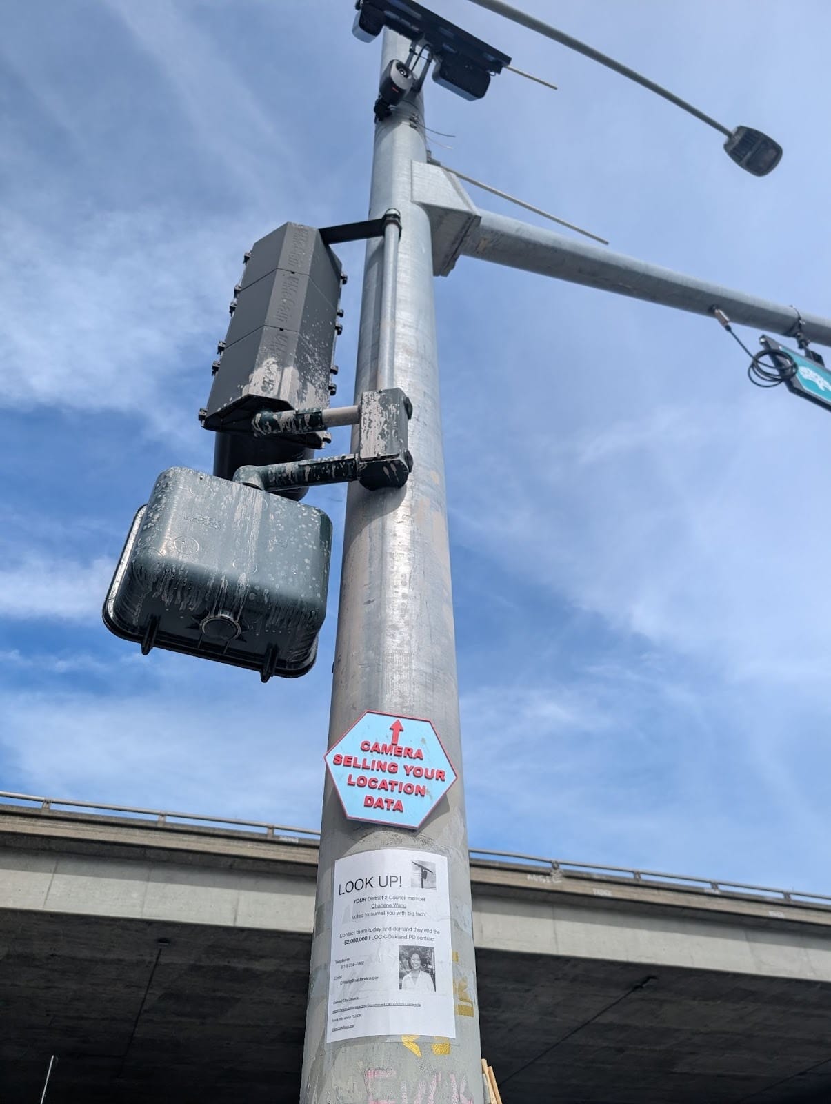 A hexagonal sign reading, "CAMERA SELLING YOUR LOCATION DATA," with an arrow pointing up toward the Flock camera. 