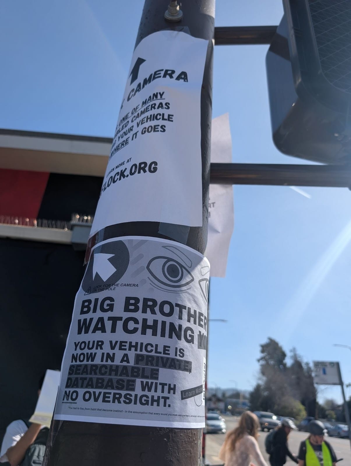 A sign on a pole, illustrated with two eyes. Text reads: "BIG BROTHER IS WATCHING."