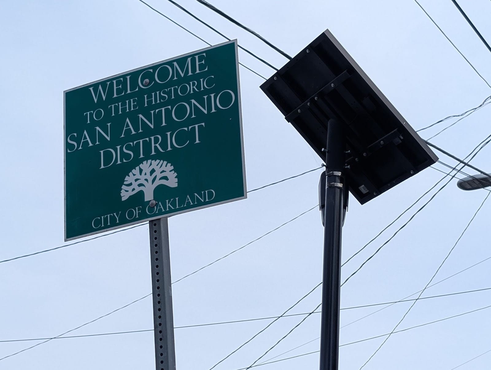 A Flock camera adjacent to a sign reading "WELCOME TO THE HISTORIC SAN ANTONIO DISTRICT" with the Oakland tree logo and "CITY OF OAKLAND" on the bottom.