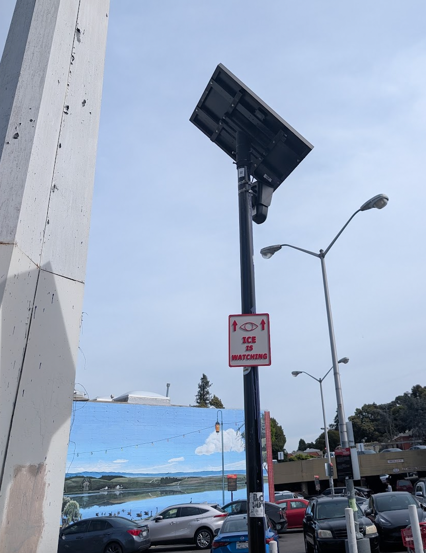 An "ICE IS WATCHING" sign hung on a pole that is topped by the Flock setup. A mural of Lake Merritt is in the background.
